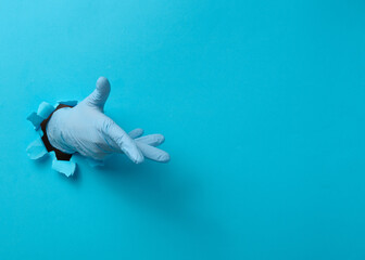 Hand in a blue medical glove is sticking out of a torn hole in a blue paper background.