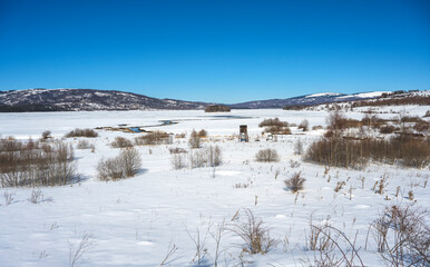 Winter landscape with a watch post and tributary River entering the frozen lake with with lake island and hills in the background background. Frozen Lake on a cold winter day.