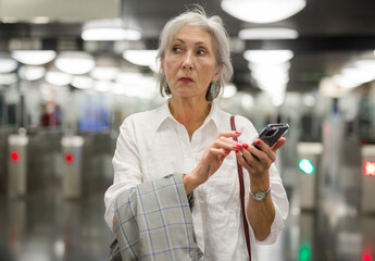 European mature woman standing near ticket barriers in metro station with her smartphone in hands.