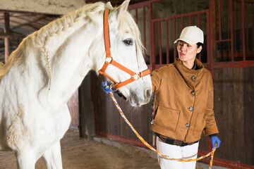 Positive Asian female stable worker leading white horse by bridle in barn. Equestrian business concept..