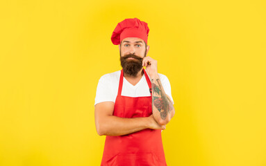 Man in red apron and toque twirling moustache with puzzled look yellow background, cook