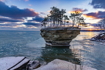 The sun begins to rise over Lake Huron and illuminate the skies and reflect off the waters surface. A rock formation known as Turnip Rock stands tall, amongst the pancake ice that's beginning to form 