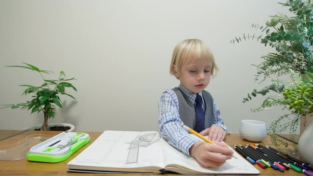 5 years old caucasian boy dressed very smart does homework in a big notebook drawing with pencil. Rulers, alidad and colorful pencils are also on the table