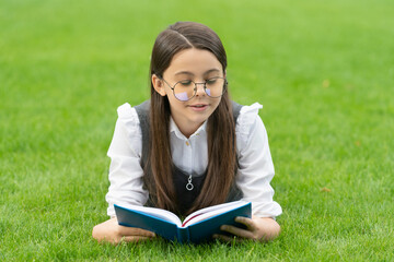 Fototapeta premium Serious schoolgirl reading book lying on grass, reading. Teen schoolgirl reading outdoors