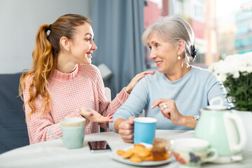 Two positive women, grandmother and her adult granddaughter, sitting at table and having conversation while drinking tea.