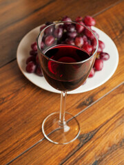 Glass of fine red wine and bunch of fresh grapes on a white plate and wooden table.