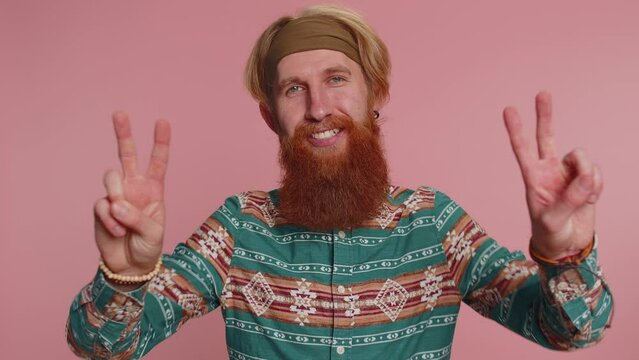 Bearded hippie man in pattern shirt showing victory V sign, hoping for success and win, doing peace gesture, smiling with kind optimistic expression. Young redhead hipster guy boy on pink background