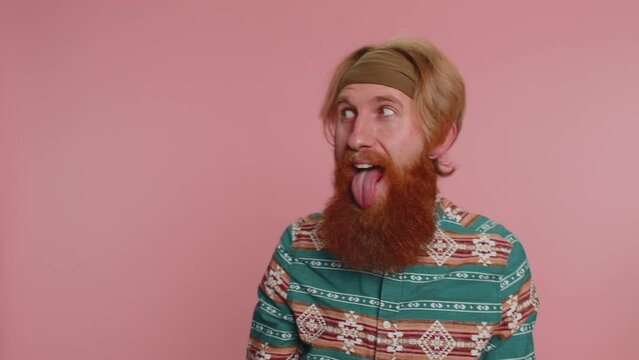 Funny joyful sincere redhead hippie man in pattern shirt making playful silly facial expressions and grimacing, fooling around showing tongue. Young handsome hipster guy isolated on pink background
