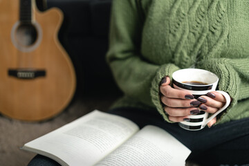 Cozy winter with woman holding coffee cup while reading a book at home. Acoustic guitar as...