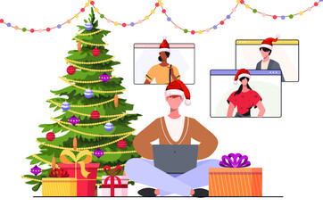 Christmas online party. A young modern man in a New Year's hat sits near the Christmas tree with a laptop in his hands celebrating New Year together with friends in a video chat.