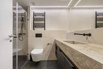 Contemporary design toilet with a large mirror that covers the wall and a marble sink with a wooden cabinet below, hanging toilet, shower with glass screen and black taps