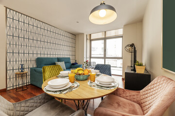 Living-dining room of a vacation rental house with breakfast service at a circular wooden table with fresh fruits and juices surrounded by chairs upholstered in different colored velvets