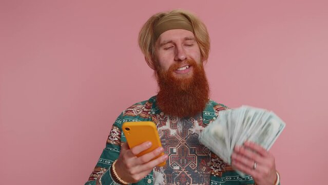Happy hippie tourist man looking smartphone display sincerely rejoicing win, receiving money dollar cash banknotes, success lottery luck. Young bearded hipster guy isolated on pink studio background