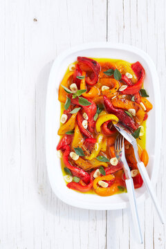 Marinated, roasted peppers with garlic, herbs and pepper with serving spoon and fork on a white wood table, studio shot on white background