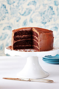 Chocolate layer cake with a few slices out on a white cake stand with a white and blue patterned background.