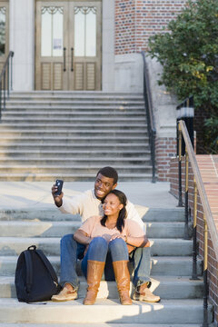 Young couple sitting together outdoors on college campus steps, taking selfie with smartphone, Florida, USA