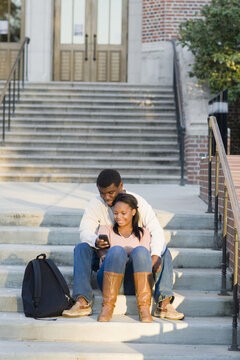 Young couple sitting together outdoors on college campus steps, looking at smartphone, Florida, USA