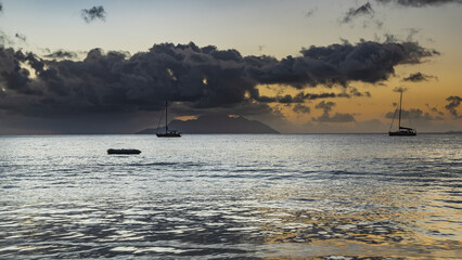 Fototapeta premium Evening twilight in the tropics. Boats are visible in the calm ocean. The sky is highlighted in orange. The island on the horizon is hidden in picturesque clouds. Seychelles. Mahe