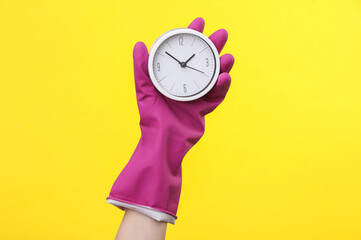 Hand in purple rubber cleaning glove holding alarm clock on a yellow background. House cleaning and housekeeping concept