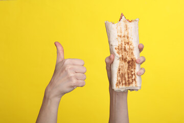Woman's hand holds an appetizing bitten shawarma and shows a thumbs up on a yellow background