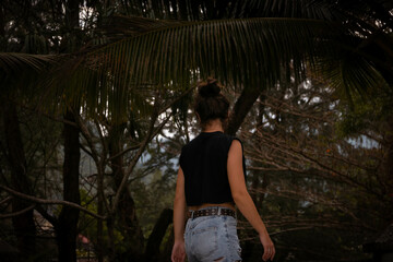 A girl walks near the beach under palm trees in denim shorts and a black T-shirt. Dark photo processing