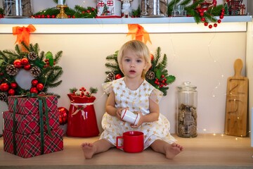 Christmas kitchen girl. Portrait of a charming little girl 2 years old, she sits on a table in a white dress in a decorated kitchen for christmas. Merry Christmas and New Year concept.