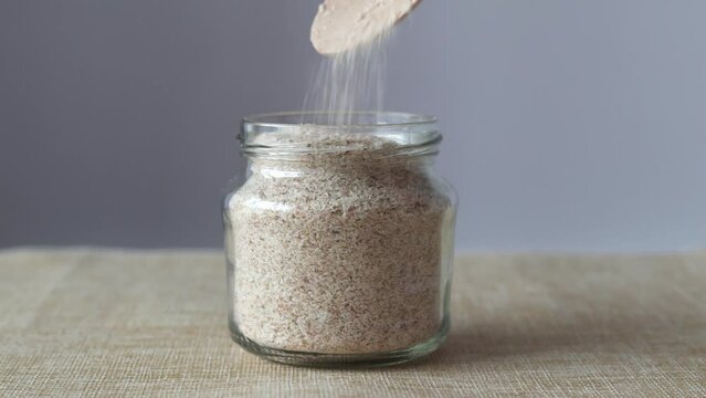 Psyllium is a plantain husk. A glass jar with a wooden spoon from which Psyllium sprinkles on a beige and gray background.