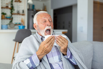 Mature man feeling sick with cold and fever at home, ill with flu disease sitting on the sofa with ice pack on his head