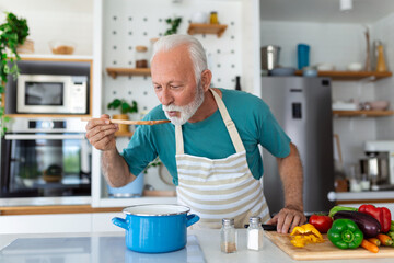Happy retired senior man cooking in kitchen. Retirement, hobby people concept. Portrait of smiling senior man holding spoon to taste food
