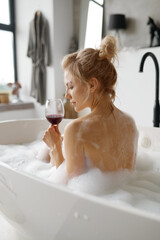 Sensual relaxed woman drinking red wine while taking bath with foam