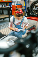 Concentrated female mechanic with clipboard reviewing documentation of motorcycle on garage