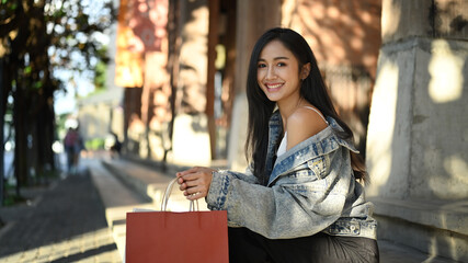 Elegant young woman holding shopping bags sitting at urban stairs with leaves shadow and sunlight 