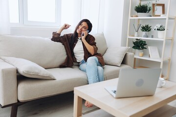 Obraz premium Woman blogger with phone lying at home and talking on the phone on sofa with laptop and working freelancer online, smiling with teeth