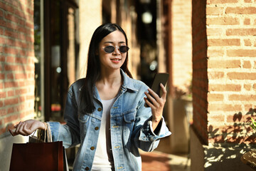 Beautiful young woman carrying shopping bags and using smart phone at outdoor on sunny day