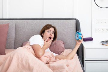 Young attractive woman waking up in the bedroom thanks to alarm clock. Discontented girl lying in...
