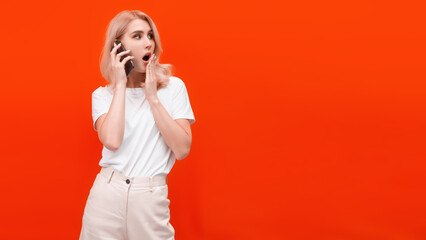 blonde woman stands on an orange background speaks on the phone and expresses the emotion of surprise
