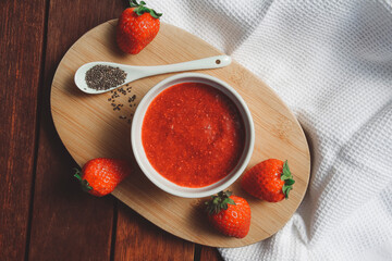 Homemade strawberry jam in a white ceramic bowl with chia seeds. Photographed on a wooden table with a white cloth in the background.