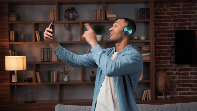 African american man dancing near sofa with smartphone in his hands listening to music shooting video for social media in headphones. Attractive cheerful guy having fun dance indoors in leisure time. 