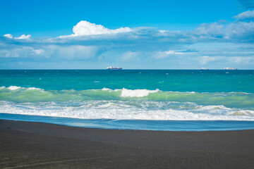 Sea waves breaking on a black sand of an empty beach. Distant cargo ship sailing at the horizon