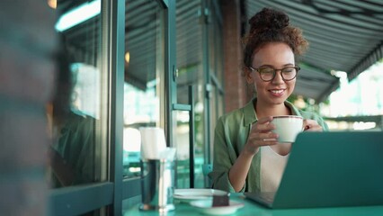Smiling African woman wearing eyeglasses drinking tea and watching something on laptop in cafe - Powered by Adobe