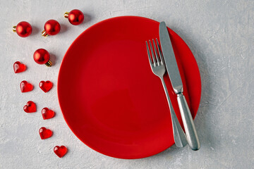 An empty red ceramic plate with cutlery surrounded by red christmas balls and hearts on a light concrete table - Powered by Adobe