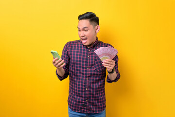 Surprised young Asian man using mobile phone and holding money banknotes isolated over yellow background. Fast credits concept