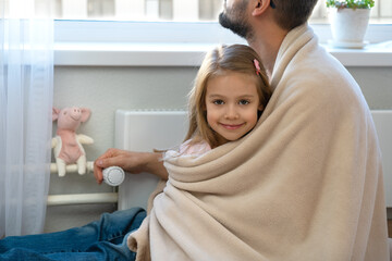 Little daughter sits with father in plaid near heating radiator with thermostat regulator in house....