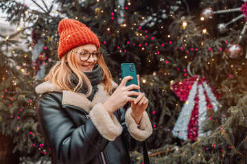 Happy blonde woman takes a picture of a European Christmas market with a smartphone. The girl enjoys the winter holidays season, visits the outdoor Christmas market, takes pictures on a mobile phone.