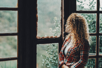 One woman standing alone at home looking outside the windows at nature background view. Female people in relax and indoor leisure activity. Side view of young lady inside chalet apartment vacation