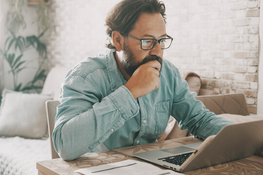 One young mature man working on laptop at home on the table in alternative workplace office. Concept of apartment and people in business job. Single male using computer and internet technology