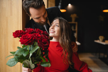 Fototapeta bearded man in suit holding bouquet of red roses near joyful girlfriend on valentines day obraz