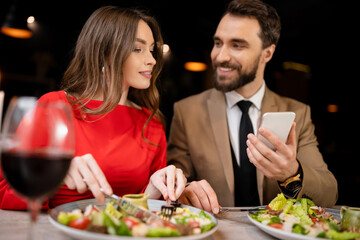 bearded man using smartphone near happy girlfriend in restaurant during celebration on valentines day