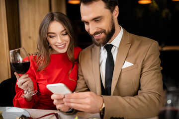 bearded man using smartphone near happy girlfriend with glass of wine in restaurant
