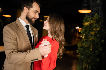 side view of cheerful man and woman in festive attire holding hands and looking at each other on valentines day